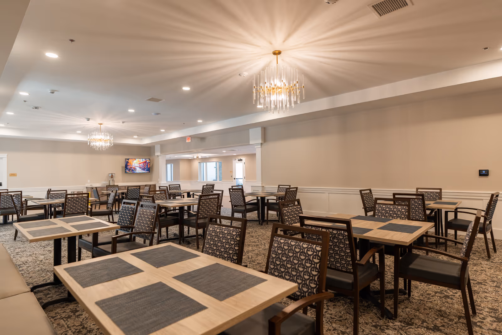 Spacious senior dining room with multiple tables and patterned chairs, chandeliers, and a TV on the far wall.