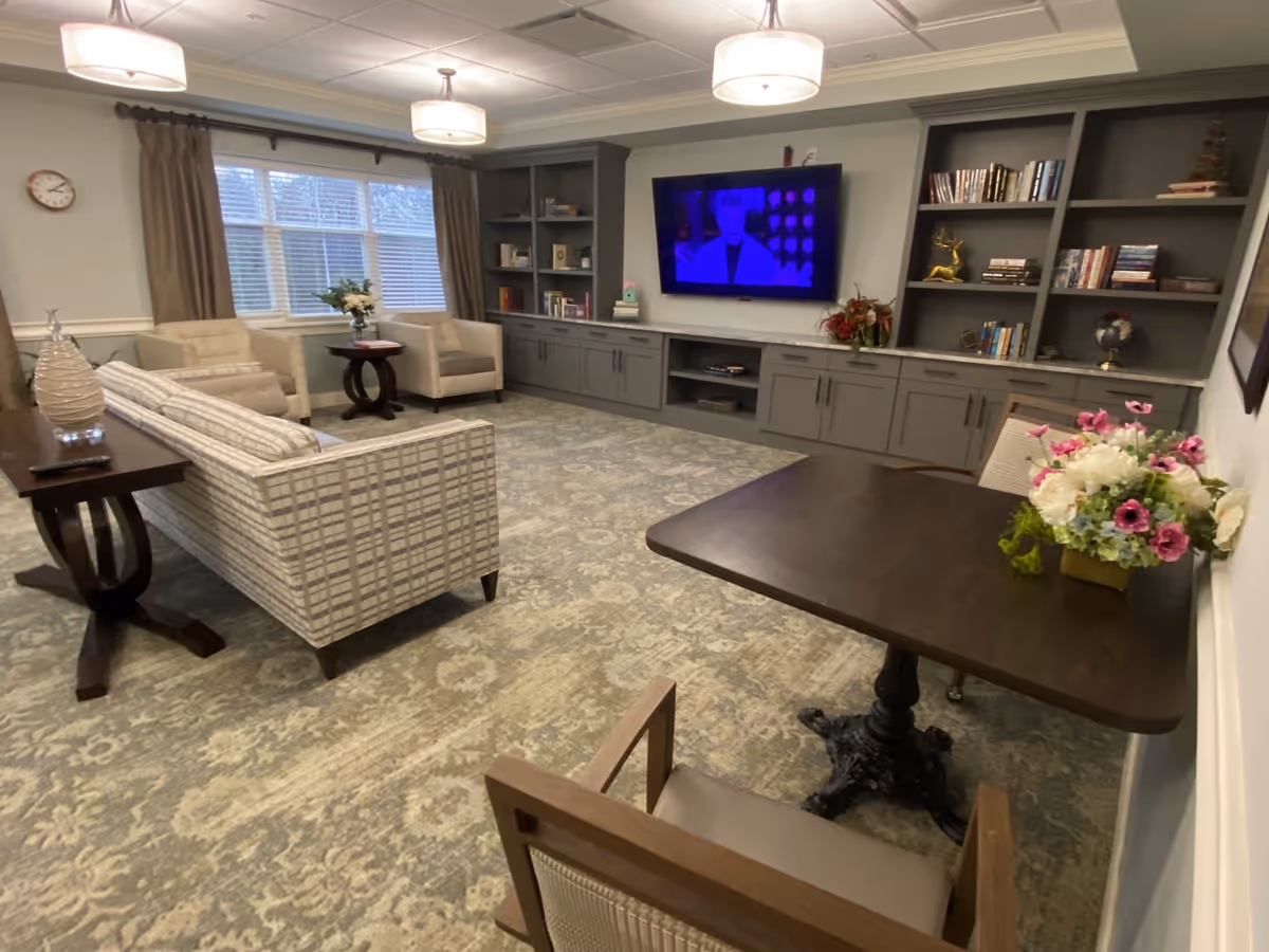 A cozy living room area with a patterned sofa, two armchairs, a small round table with a flower vase, a dark wooden table with a floral arrangement, built-in gray bookshelves filled with books and decorative items, and a wall-mounted flat-screen TV. The room has large windows with brown curtains and ceiling lights.