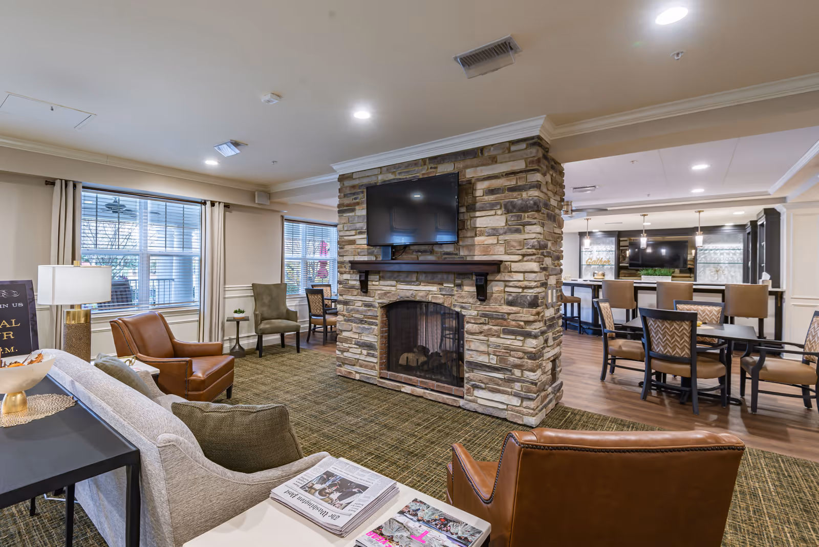 A cozy senior living facility common area featuring a stone fireplace with a mounted flat-screen TV above it. The room includes comfortable seating such as leather and fabric armchairs, a sofa, and dining tables with chairs. Large windows with curtains allow natural light to fill the space, and the area has a warm, inviting atmosphere with carpet and wood flooring.