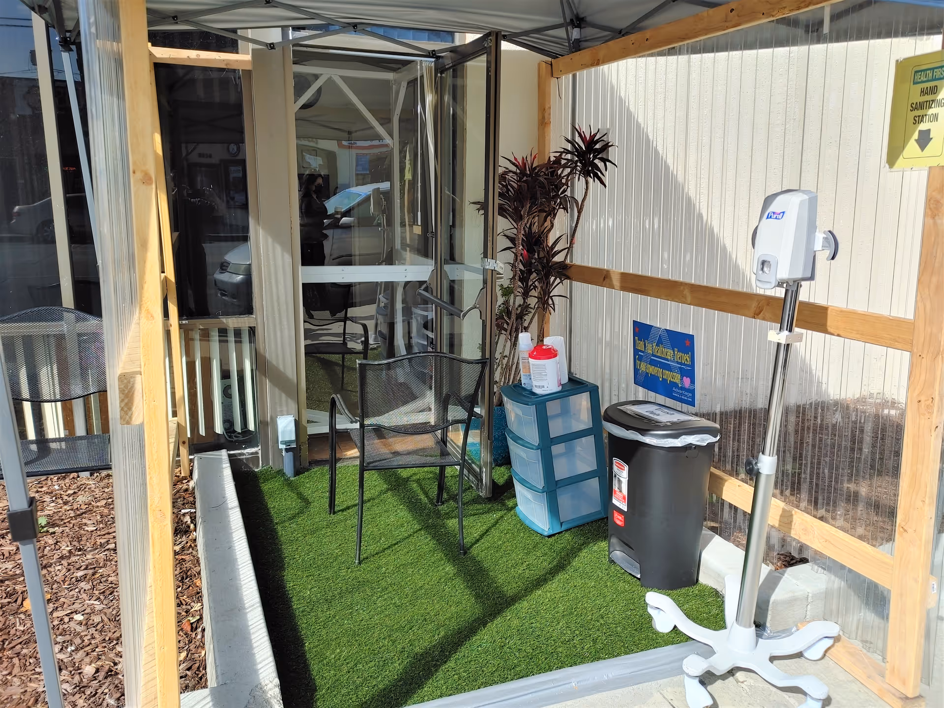 A small enclosed outdoor area with artificial grass flooring, a black metal chair, a plastic drawer unit with cleaning supplies on top, a trash can, a hand sanitizing station on a stand, and a potted plant. The enclosure has transparent walls and a door leading inside a building.