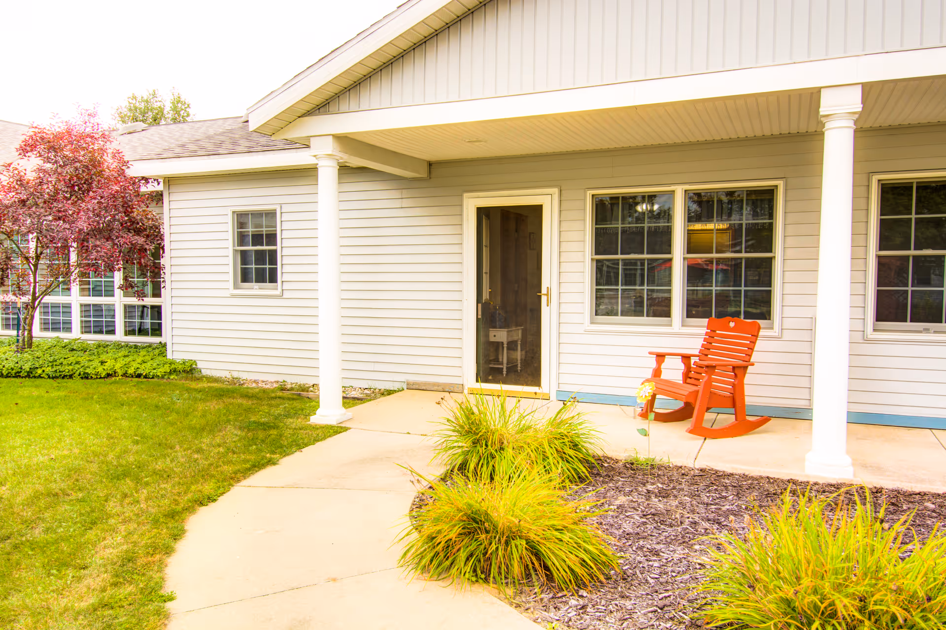 Exterior view of a single-story building with light gray siding, white columns, and a covered porch. There is a red rocking chair on the porch and green grass and plants surrounding the walkway leading to the door.