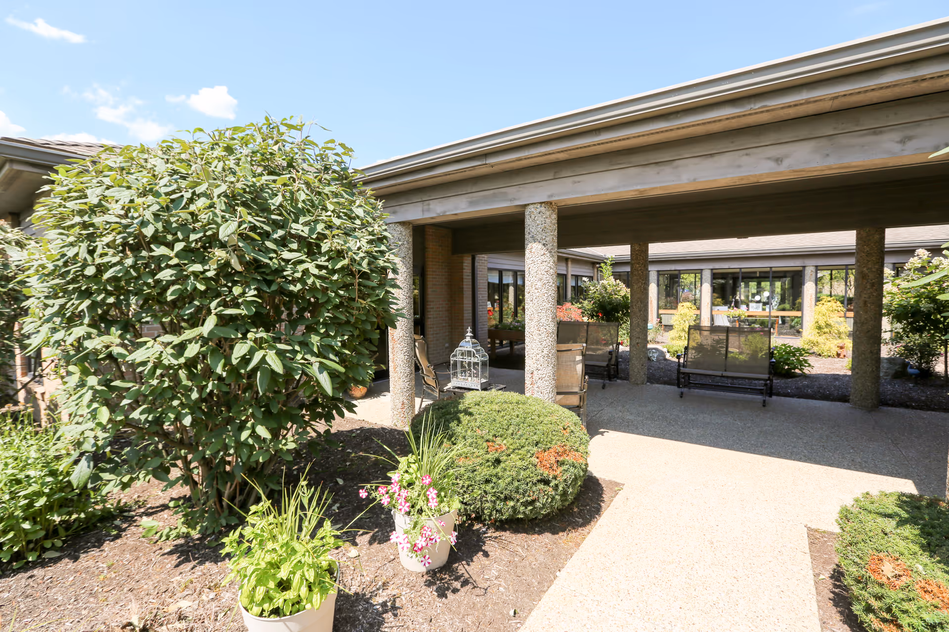 Outdoor courtyard area at Brookdale Medina South featuring a covered walkway supported by stone columns, surrounded by green shrubs, potted plants with flowers, and garden beds under a clear blue sky.