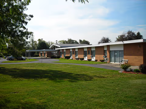 Exterior view of a single-story brick building with multiple windows and a covered entrance, surrounded by a well-maintained lawn and trees under a partly cloudy sky.