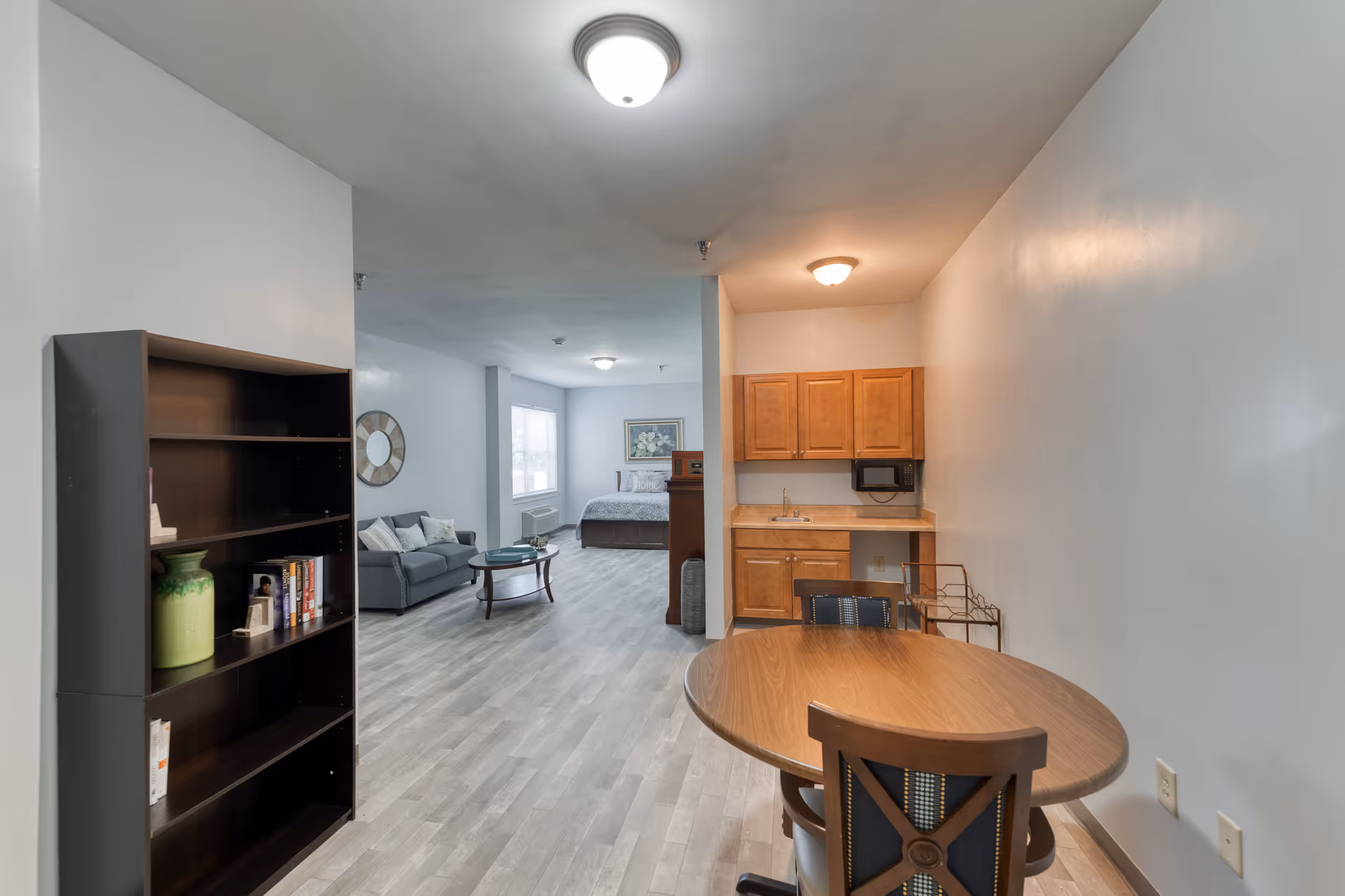 Interior view of a senior living facility apartment with a small kitchenette featuring wooden cabinets, a microwave, and a sink. In the foreground, there is a round wooden dining table with chairs. Further back, there is a living area with a gray sofa, a round coffee table, and a bookshelf on the left. At the far end, a bed is visible near a window with natural light coming in. The walls are painted light gray and the floor has a light wood finish.
