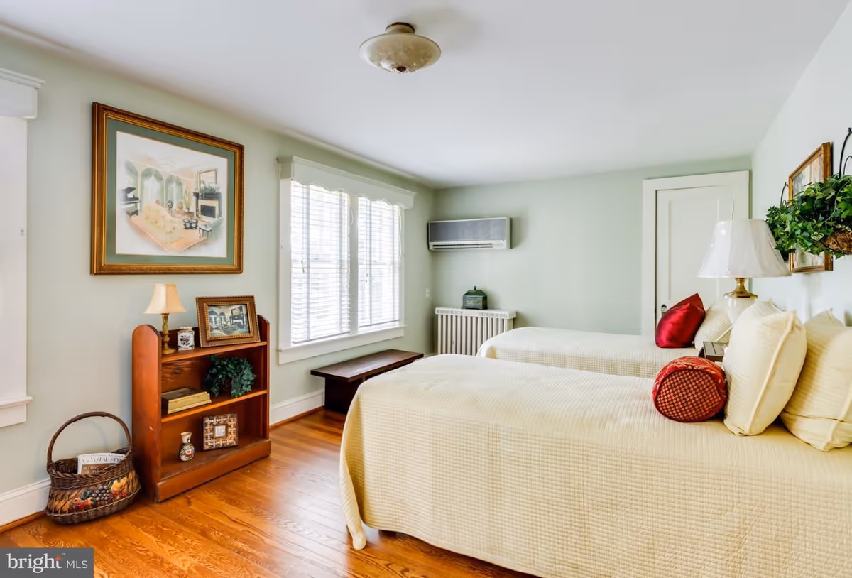 A bright bedroom with two twin beds covered in cream-colored quilts and red accent pillows. The room has light green walls, a wooden floor, a window with white blinds, a small wooden bench under the window, and a wooden bookshelf with decorative items and framed pictures. A wall-mounted air conditioning unit is above a radiator, and there is a white door on the far wall.