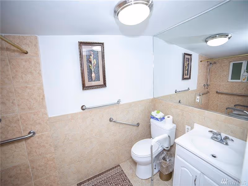 A small bathroom with beige tiled walls and floor, featuring a white toilet with a grab bar, a white sink with a cabinet, a large mirror above the sink, a framed floral picture on the wall, and multiple grab bars for accessibility. There is a patterned floor mat in front of the toilet and a ceiling light fixture.