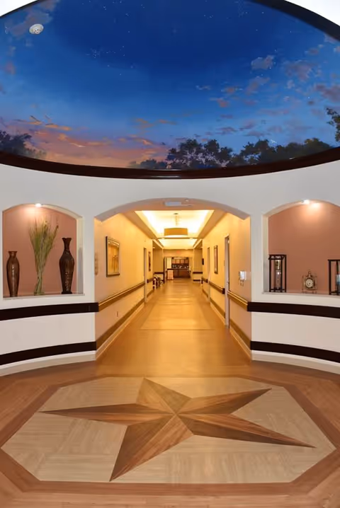 A well-lit hallway in a senior living facility with a decorative star pattern on the floor and a ceiling mural depicting a twilight sky with clouds and trees. The hallway has arched alcoves on either side containing decorative vases and clocks.