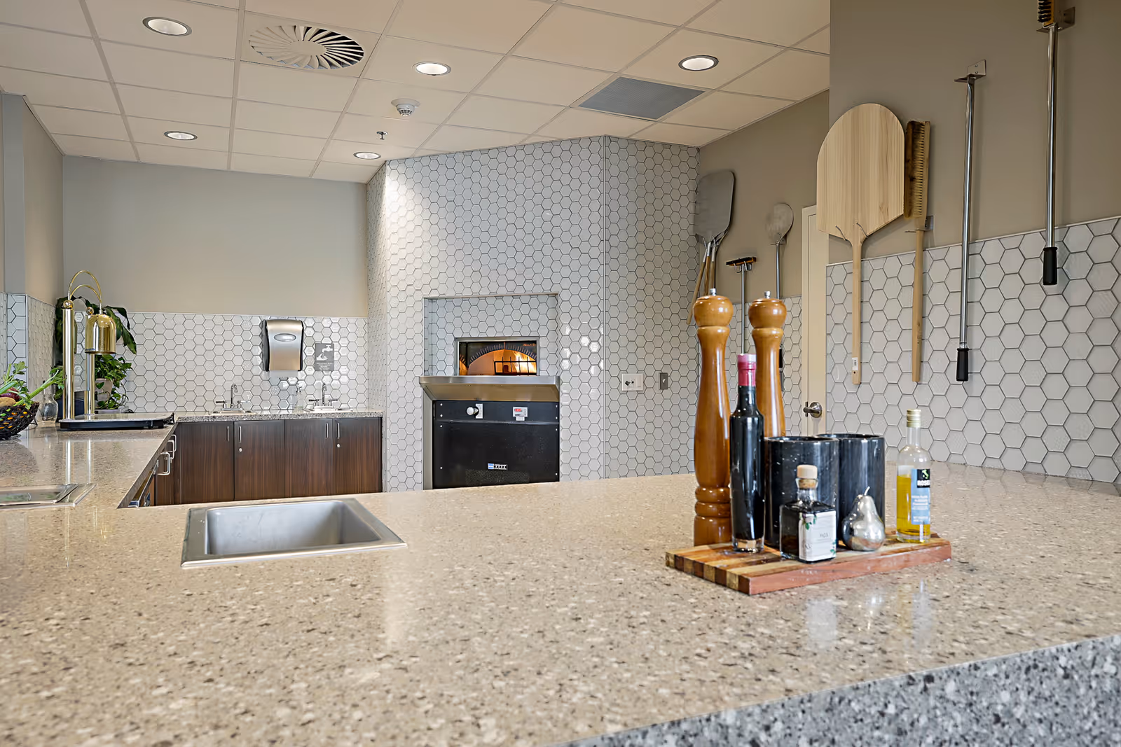 Modern communal kitchen with a large stone countertop, sink, pizza oven, and cooking utensils and condiments on display.