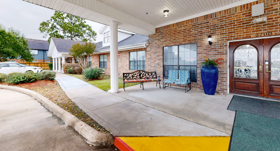 Entrance area of Cambridge Square Assisted Living featuring a covered walkway with benches, a large blue planter with greenery, brick exterior walls, and double wooden doors with decorative glass panels. There is a sidewalk leading to the entrance and some landscaping with bushes and trees nearby.