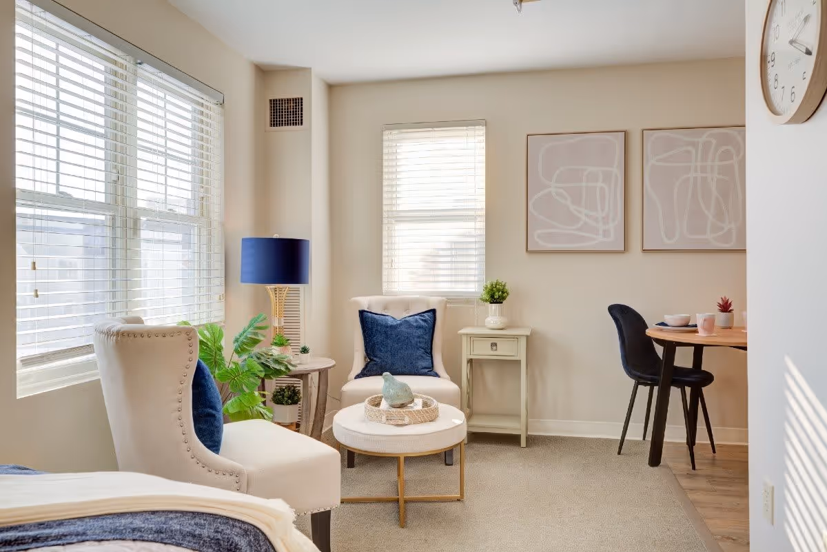 A bright and cozy living area with two beige armchairs, one with a blue pillow, a small round coffee table with decorative items, a side table with a plant, a blue lamp, and two abstract art pieces on the wall. A small dining table with two black chairs is visible in the background, along with a wall clock.