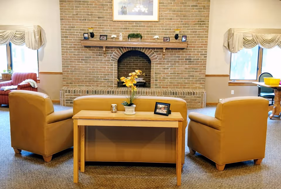 A cozy living room area with a brick fireplace as the focal point. In front of the fireplace, there is a tan leather sofa and two matching armchairs arranged around a wooden console table with a potted yellow orchid, a candle, and a framed photo. The room has carpeted flooring, two windows with beige valances, and additional seating visible in the background.
