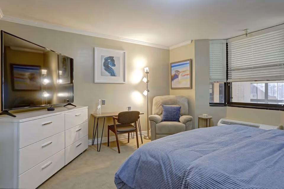 Furnished bedroom with a bed, dresser and TV, armchair, small desk, and window with blinds.