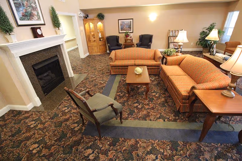 A cozy living room area with patterned carpet, two orange striped sofas, a wooden coffee table with a decorative item, a green upholstered armchair, two dark blue armchairs near a wooden display cabinet, a fireplace with a white mantel, and several table lamps providing warm lighting.
