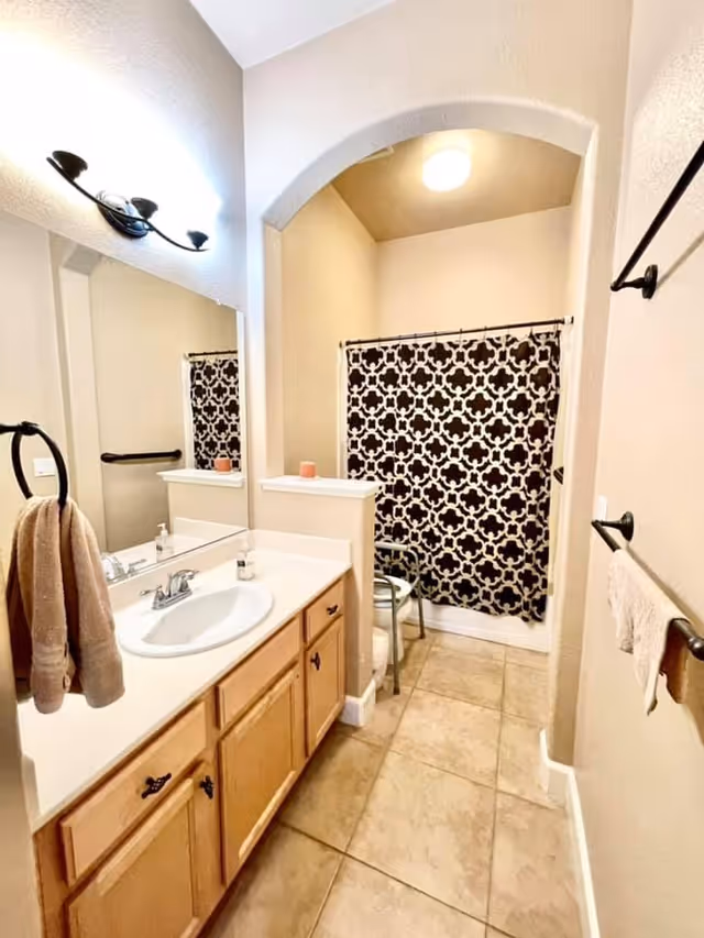 Bright bathroom with a single-sink vanity, large mirror, tiled floor and a patterned shower curtain behind an arched alcove.