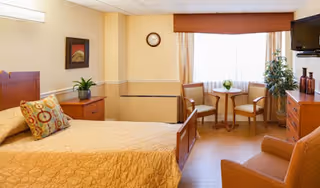 A cozy bedroom in a senior living facility with a single bed covered in a yellow quilt, a colorful pillow, a wooden nightstand with a plant, a small round table with two chairs near a window with curtains, a wall clock, a TV mounted on the wall, and a comfortable armchair.