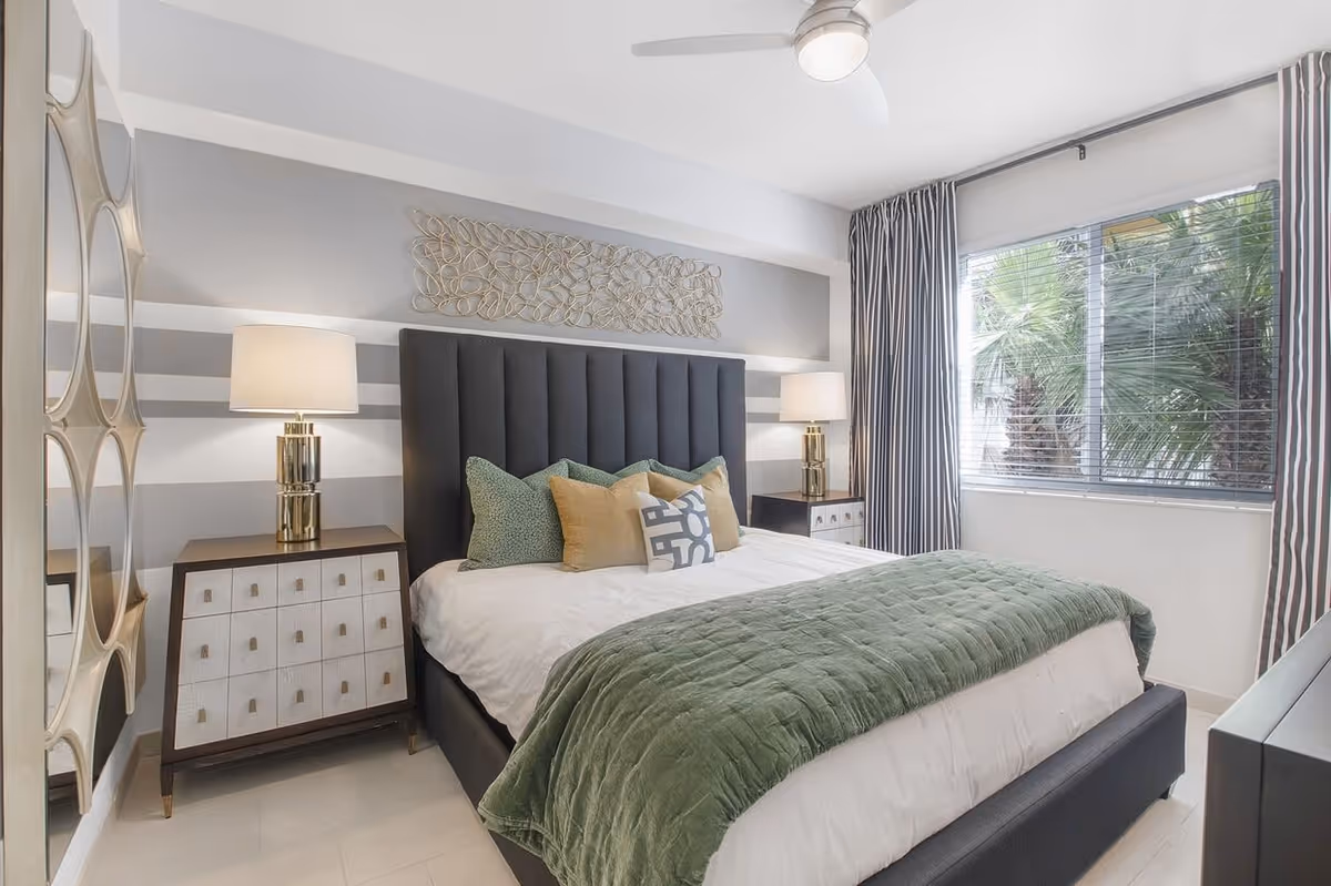 A modern bedroom with a large bed featuring a dark upholstered headboard, white bedding, and green and beige pillows. There are two matching bedside tables with lamps on each side of the bed. A decorative metal wall art piece hangs above the headboard. A large window with striped curtains lets in natural light and shows palm trees outside. A ceiling fan is mounted on the white ceiling.