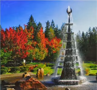 A tall, tiered water fountain in a landscaped outdoor area with vibrant red and green trees in the background under a clear blue sky.