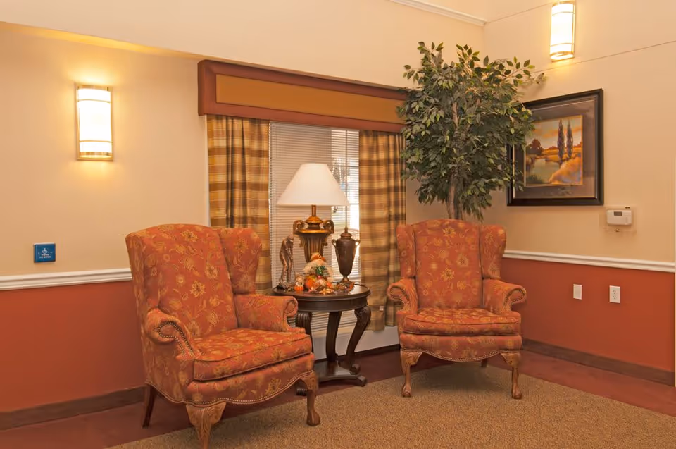 Two upholstered armchairs flank a small table with a lamp and decorations in a warmly lit sitting area.