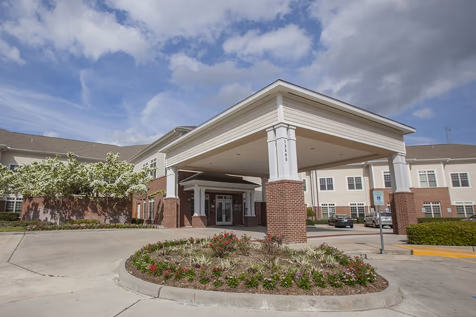 Front entrance of a senior living facility with a covered porte-cochere, driveway, and landscaped circular flower bed.