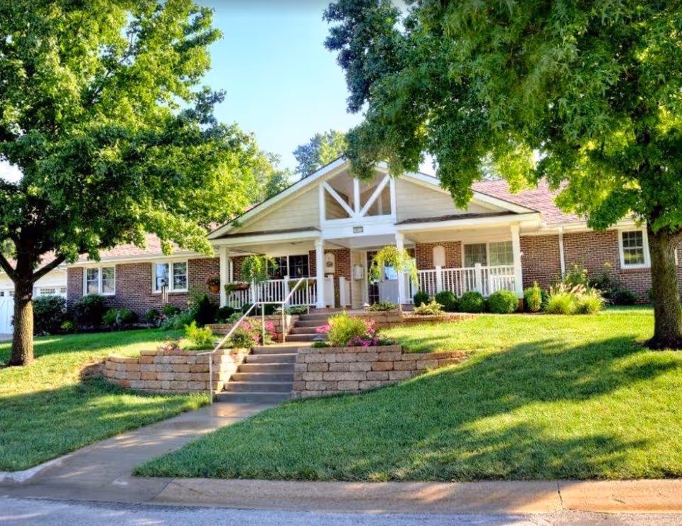 Single-story brick building with a covered front porch, steps and landscaped lawn framed by large trees.