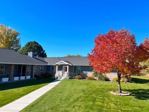 Exterior view of a single-story senior living facility building named The Heights, featuring a well-maintained green lawn, a concrete walkway leading to the entrance, and a tree with vibrant red autumn leaves under a clear blue sky.