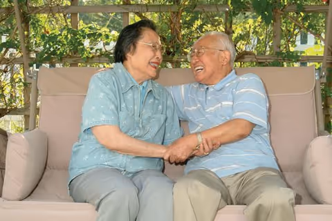 An elderly couple sitting on a cushioned outdoor bench under a wooden pergola, smiling and holding hands while looking at each other. Green foliage is visible in the background.