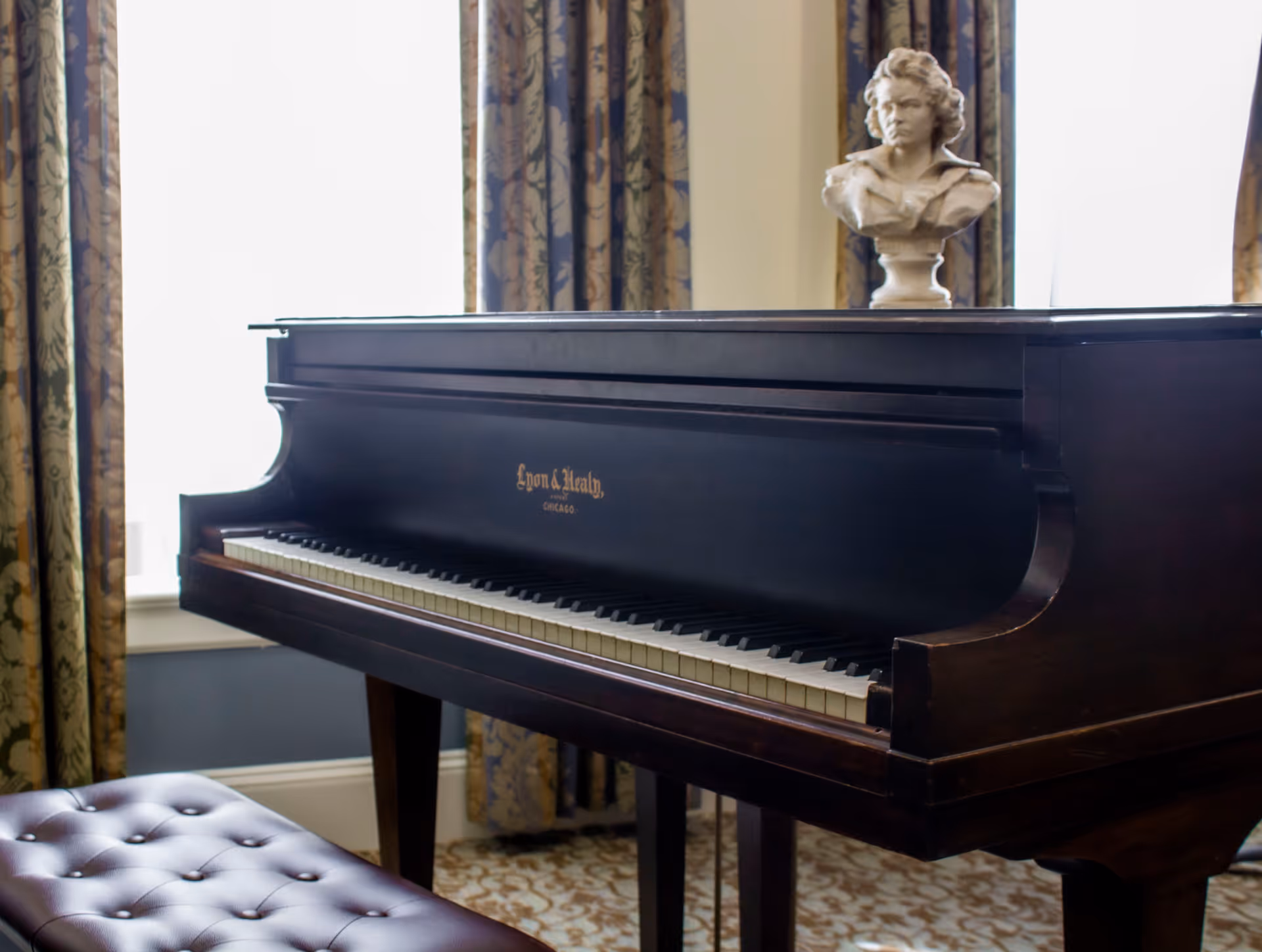 A dark wooden grand piano with a bust sculpture placed on top, situated in a room with patterned curtains and a cushioned piano bench in front.