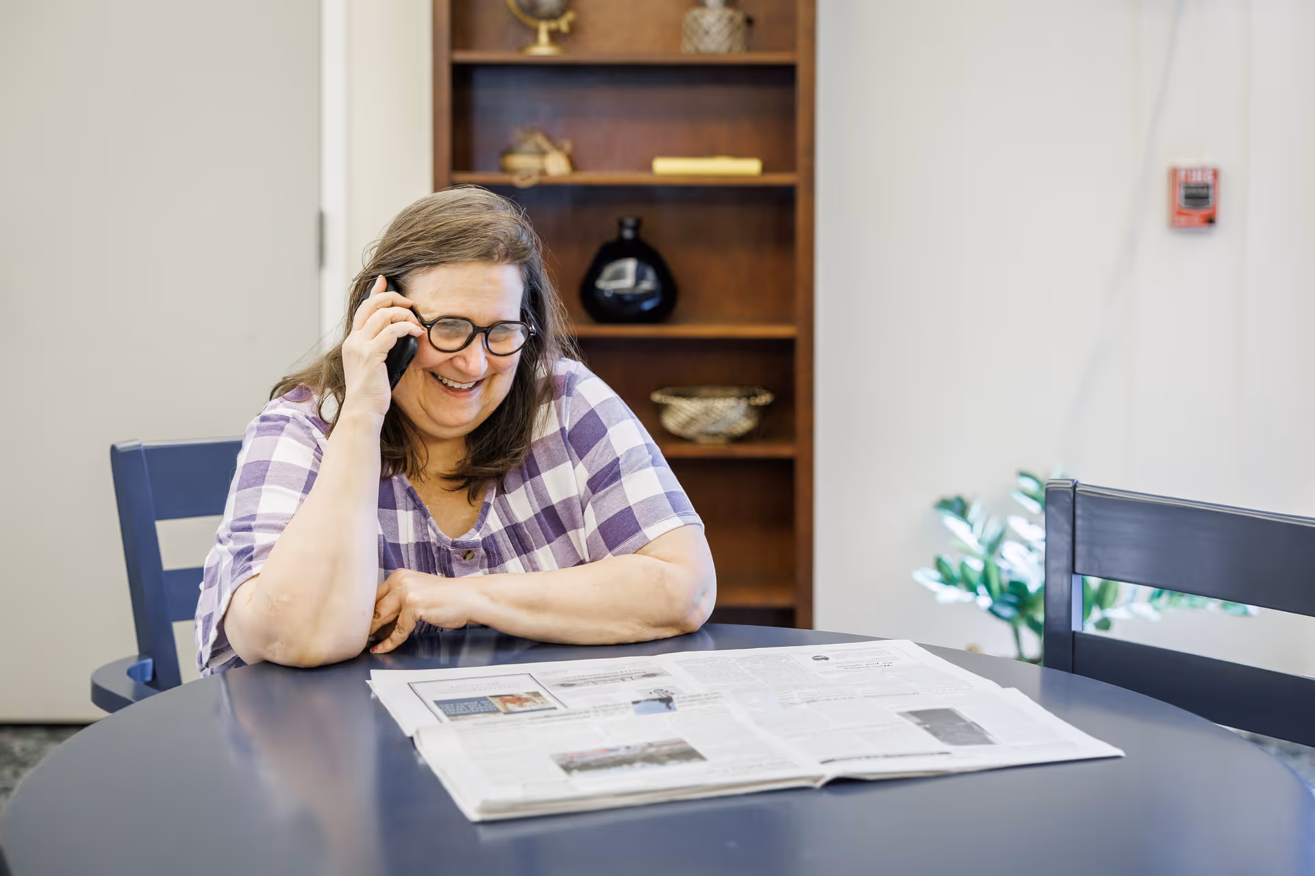 A woman with glasses and a purple checkered shirt is sitting at a round table, smiling and talking on a phone. There is an open newspaper on the table in front of her. Behind her is a wooden shelf with decorative items and a plant near the wall.