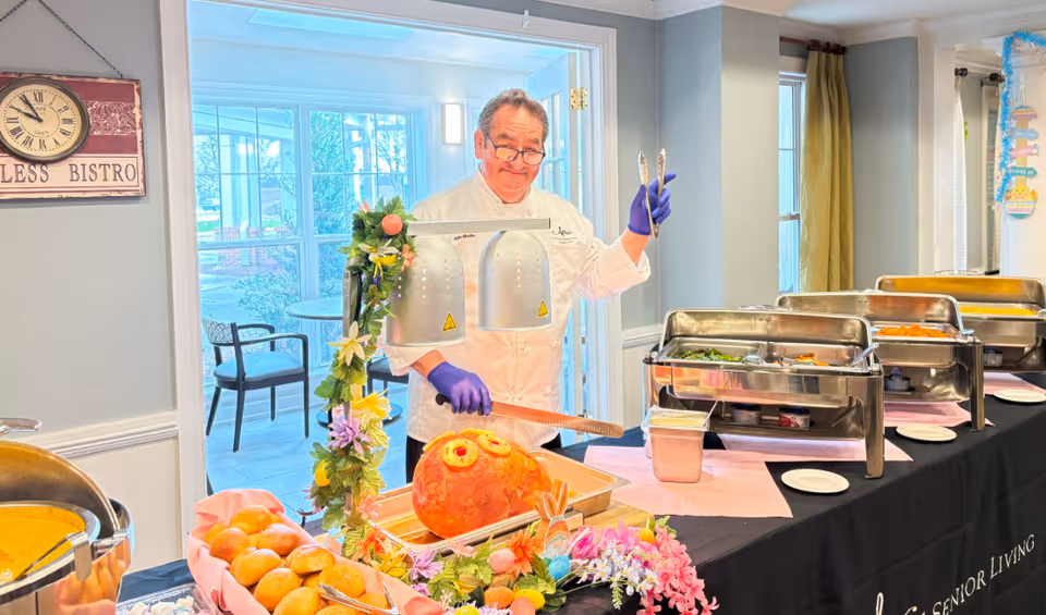 A chef wearing a white uniform and purple gloves stands behind a buffet table with various food trays and a large decorated ham. The table is adorned with flowers and Easter-themed decorations. Behind the chef, there is a bright room with large windows and chairs. A sign on the wall reads 'Endless Bistro.'