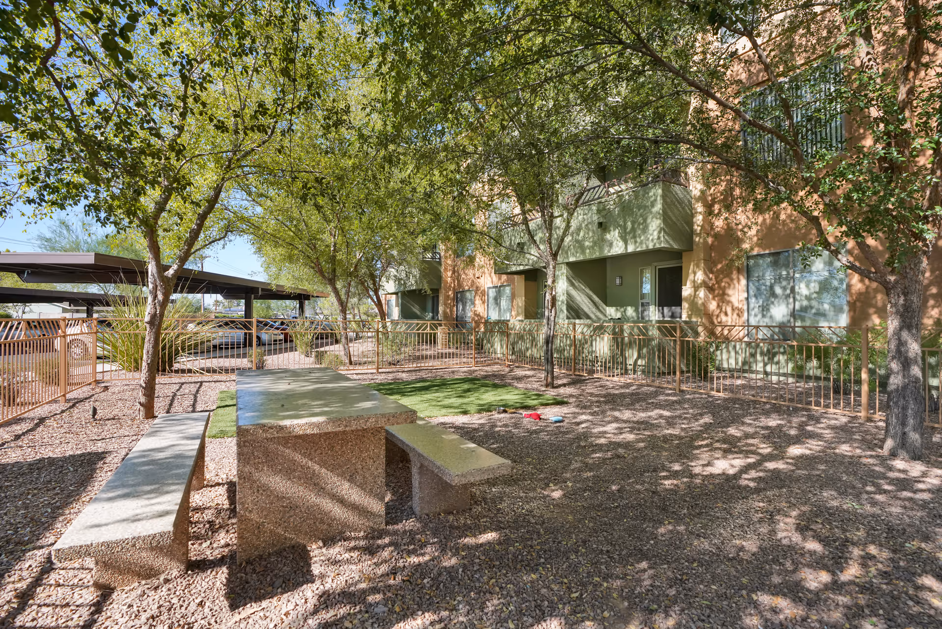 Outdoor area at McDowell Village featuring a stone picnic table with benches under trees providing shade. The area is surrounded by a metal fence, with a building and covered parking visible in the background.