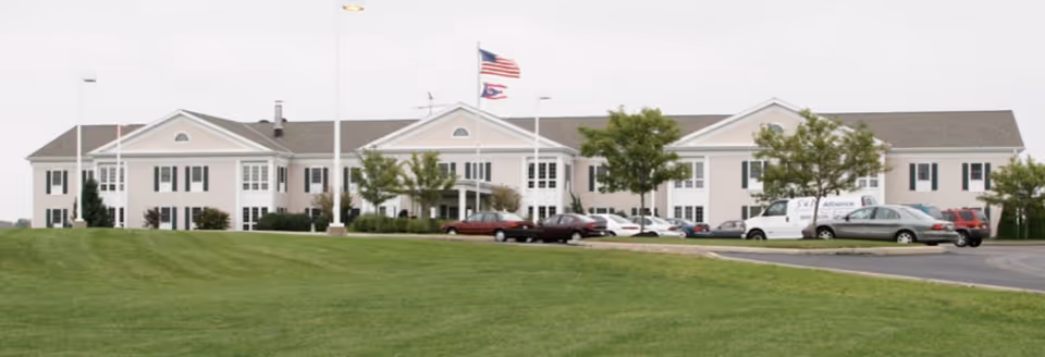 Front exterior of a two-story senior living facility with flagpoles, parked cars, and a large grassy lawn.