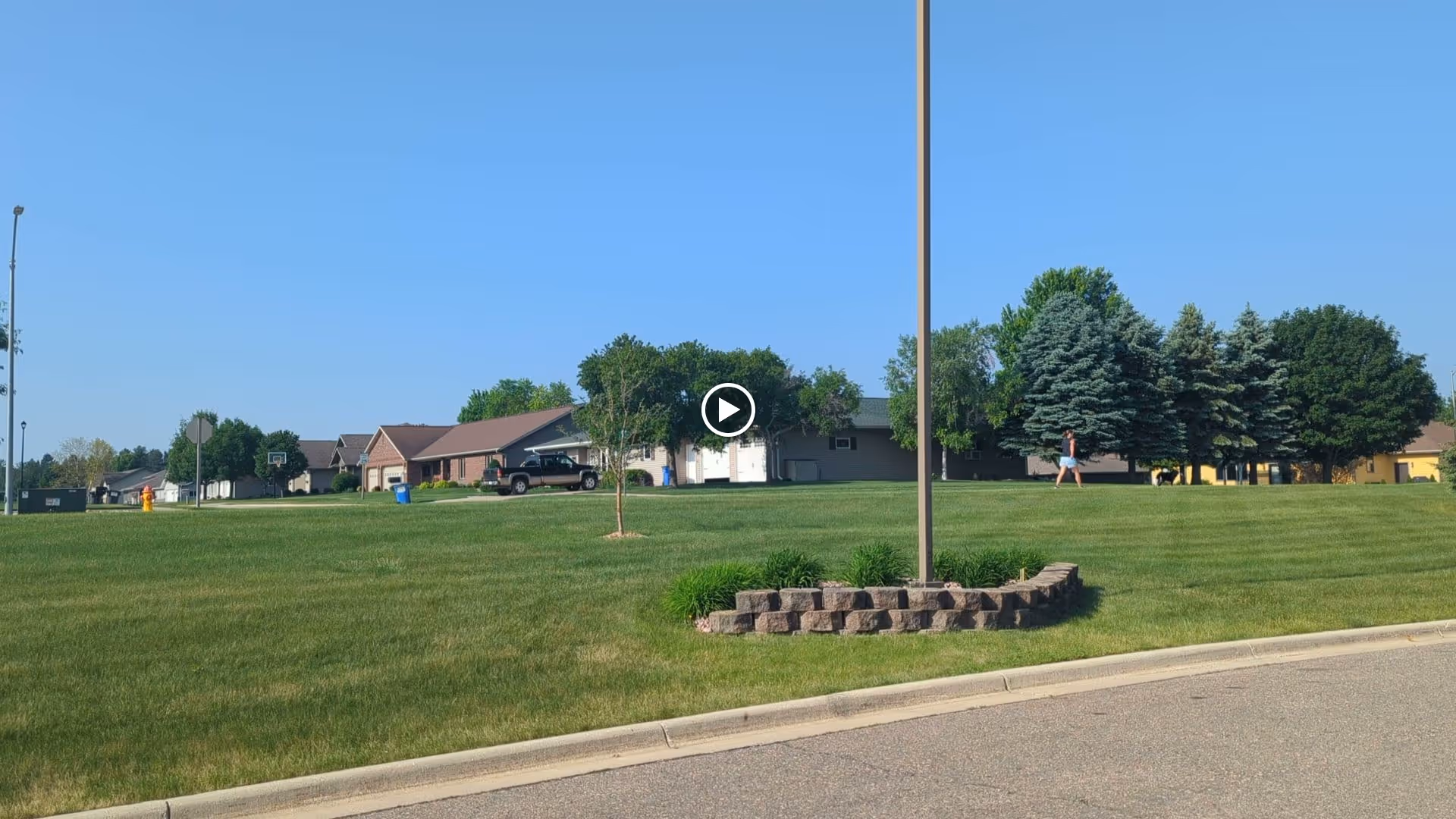 A grassy area with a small tree and a raised flower bed made of bricks surrounding a tall light pole. In the background, there are several houses, trees, and a person walking on the grass under a clear blue sky.