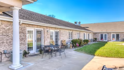 Sunny outdoor courtyard with patio tables and chairs in front of a single-story brick building with multiple French doors.