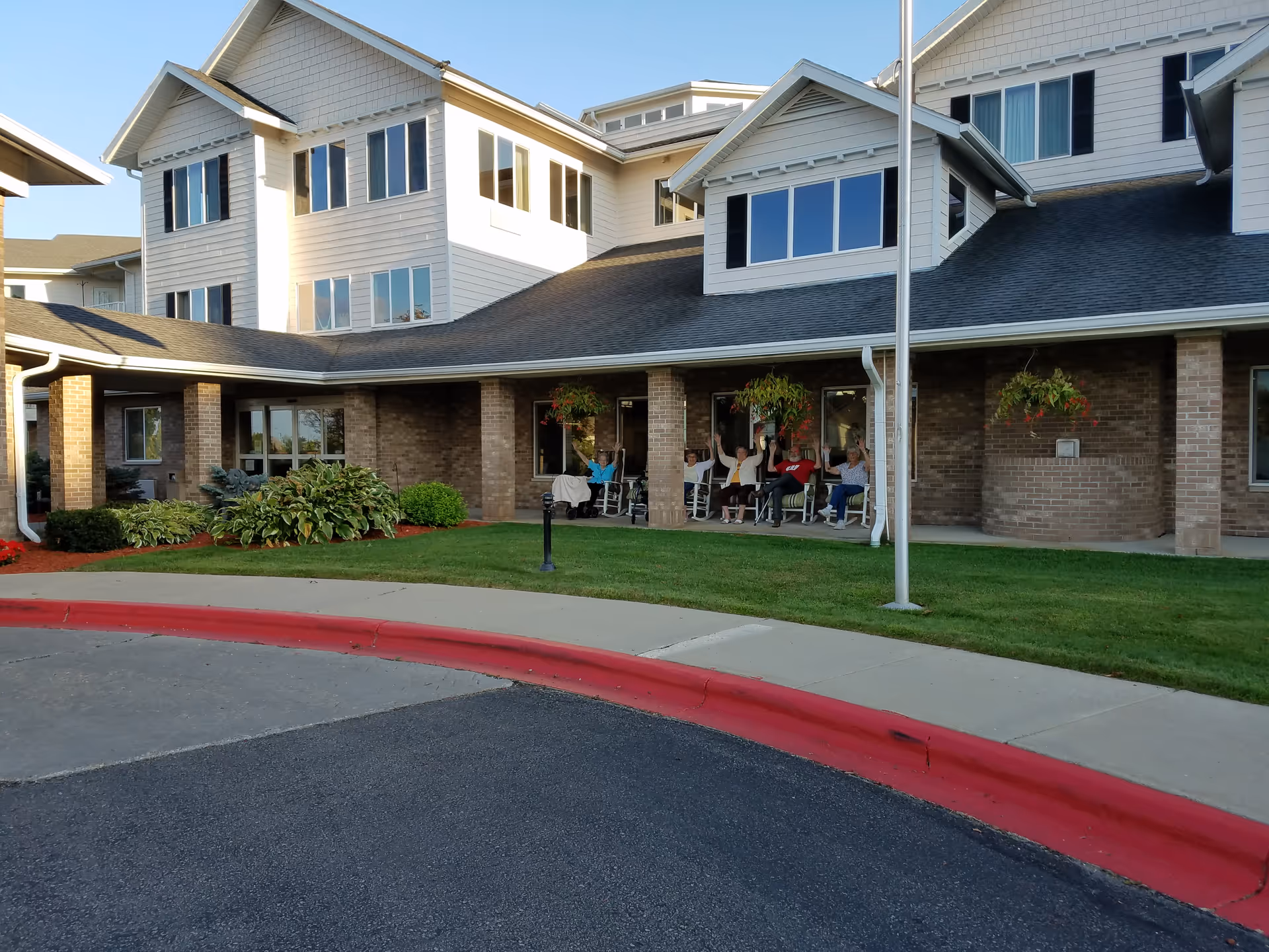 Exterior view of Middleton Crossing - Sky Active Living facility showing a multi-story building with white siding and brick columns. Several elderly people are sitting on chairs on the covered porch, waving and enjoying the outdoor space. The area is landscaped with green grass, plants, and hanging flower baskets under a clear blue sky.