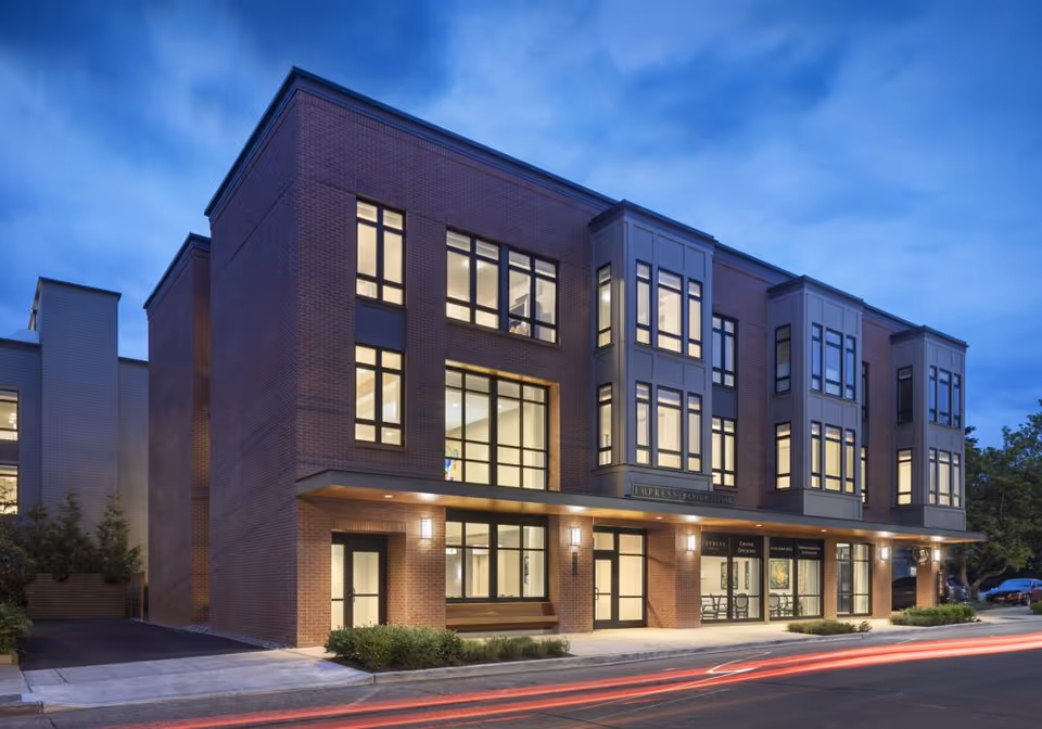 Exterior view of a modern three-story brick building with large windows illuminated from inside during dusk, identified as Empress Senior Living, with a sidewalk and some greenery in front and light trails from passing cars on the street.