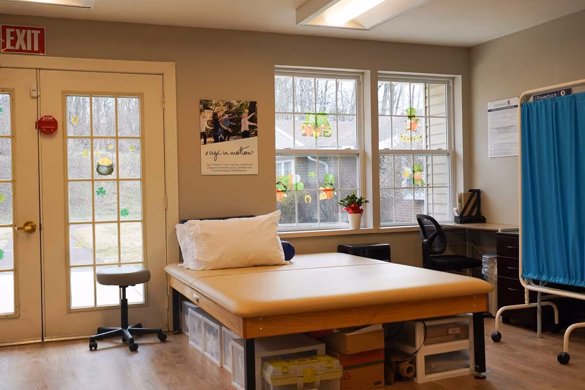 A therapy or examination room with a padded treatment table topped with a white pillow, a rolling stool, a desk with a black office chair, and a blue privacy curtain. The room has large windows decorated with St. Patrick's Day themed stickers and a glass door with an exit sign above it. There is a small potted plant on the windowsill and a poster on the wall.