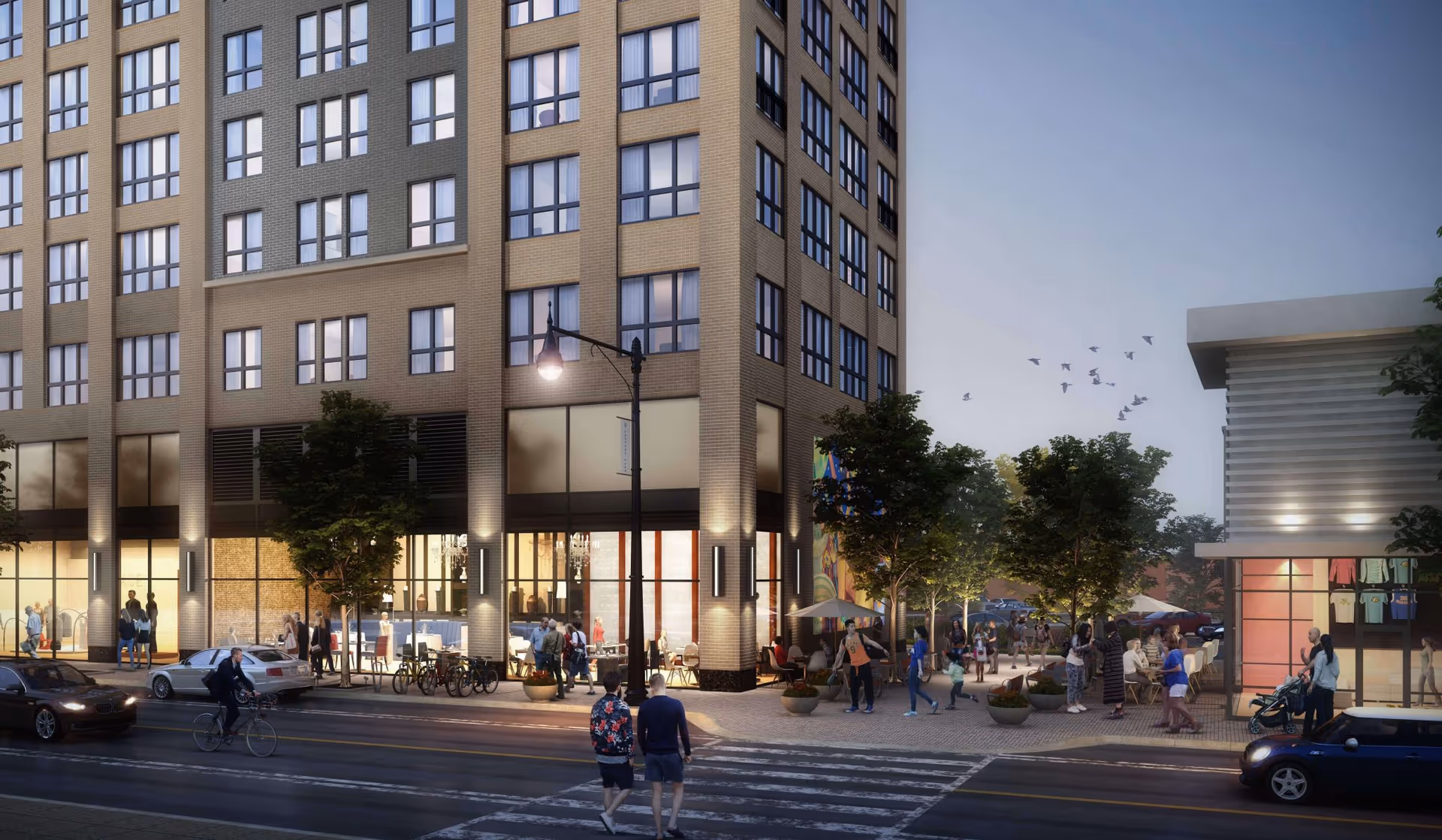 Street view of a modern multi-story building with large windows and a well-lit ground floor. People are walking on the sidewalk and crossing the street. There are trees, outdoor seating areas, and a small building with clothing displayed inside. The scene is set during dusk with a clear sky and birds flying.