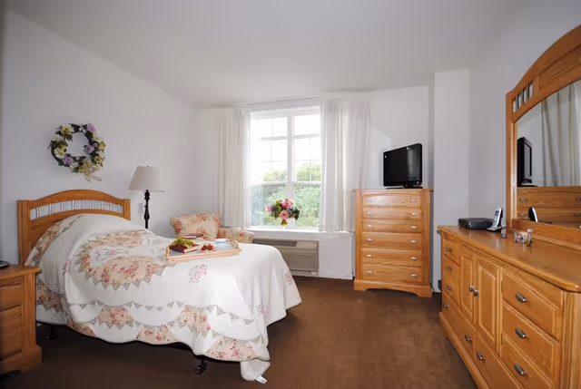 Bright bedroom with a floral-quilted bed, wooden dressers and mirror, armchair, TV and a window with curtains.