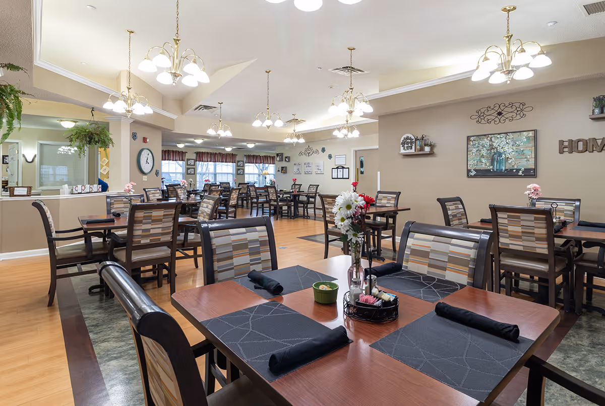 A well-lit dining room with multiple wooden tables and chairs arranged neatly. Each table has black placemats and rolled black napkins, with some tables decorated with small flower arrangements. The room features beige walls, several hanging light fixtures, and windows with curtains in the background. Wall decorations include a painting, shelves with plants, and the word 'HOME' displayed on the wall.