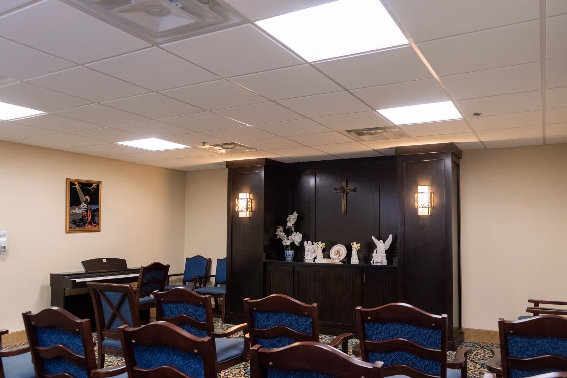 Interior room with rows of wooden chairs with blue cushions facing a dark wood cabinet adorned with religious statues and a cross on the wall above. A piano is positioned against the left wall, and a religious painting hangs above it. The room has a beige wall and ceiling with recessed lighting.