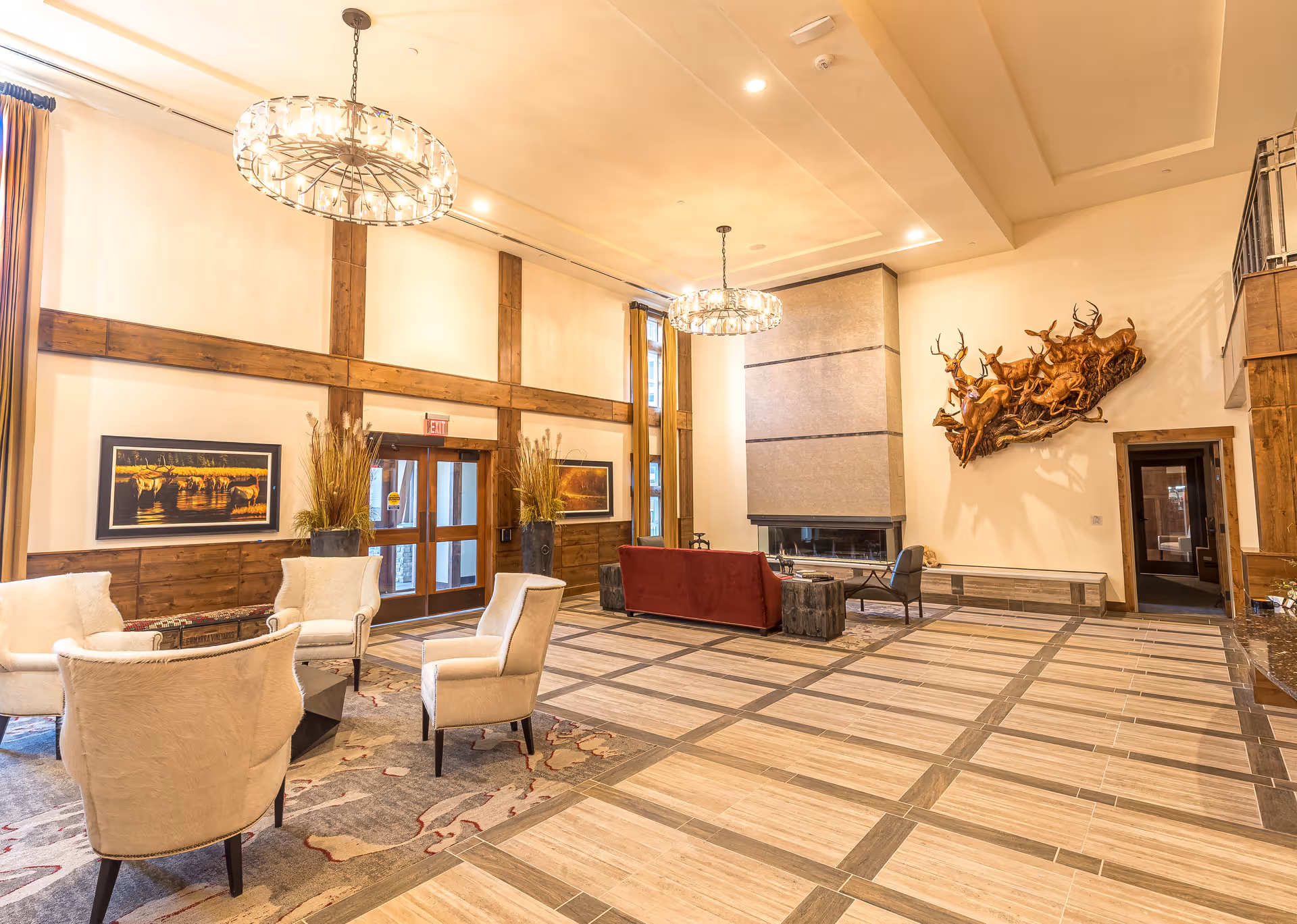 Spacious and well-lit living room area with high ceilings, featuring a modern fireplace, a red sofa, several beige armchairs arranged around a coffee table, large windows with curtains, two circular chandeliers, wooden wall accents, framed artwork, and a decorative wall sculpture of running deer.
