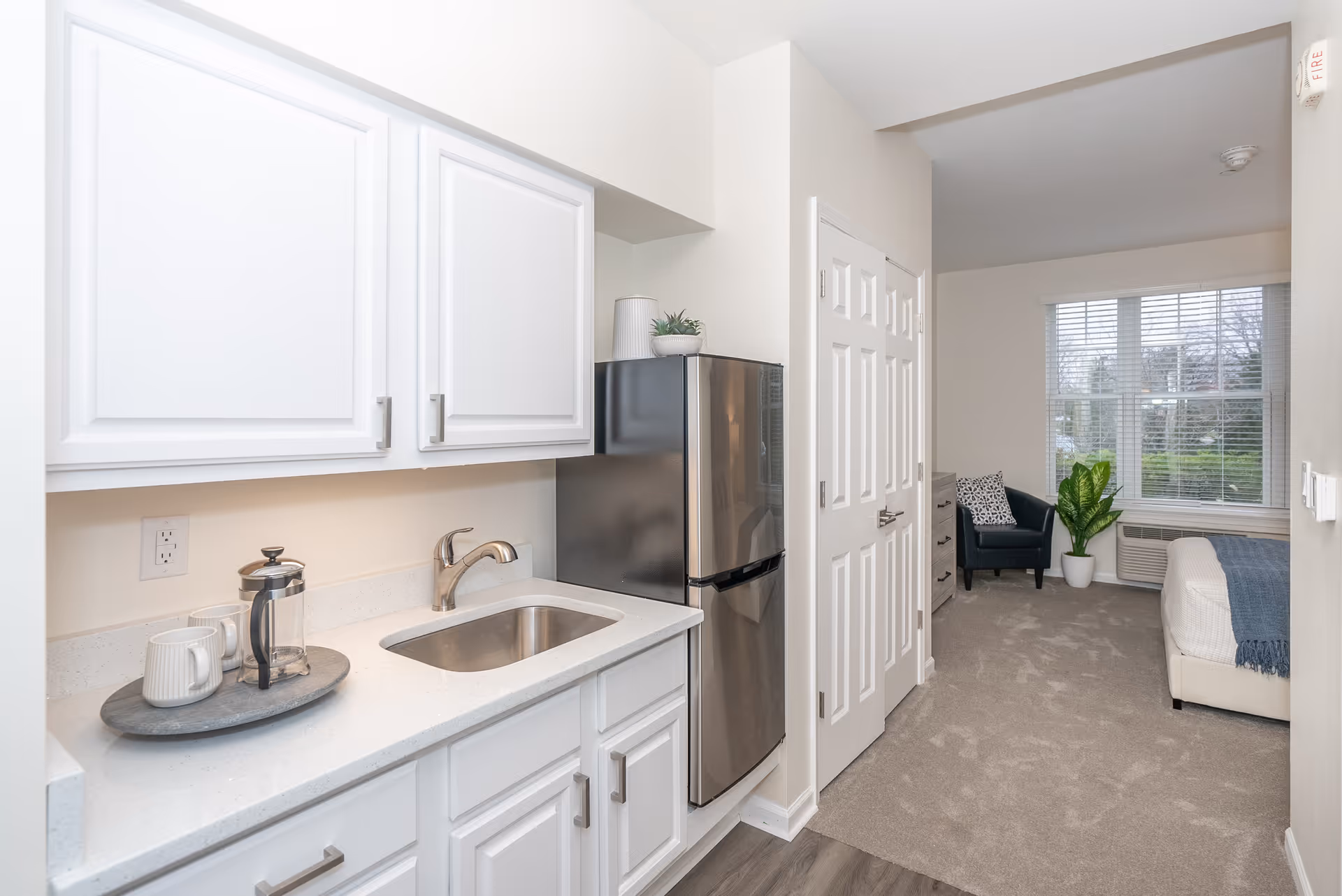 Bright kitchenette with white cabinets, a sink and stainless steel mini-fridge opening into a studio living area with a chair and window.