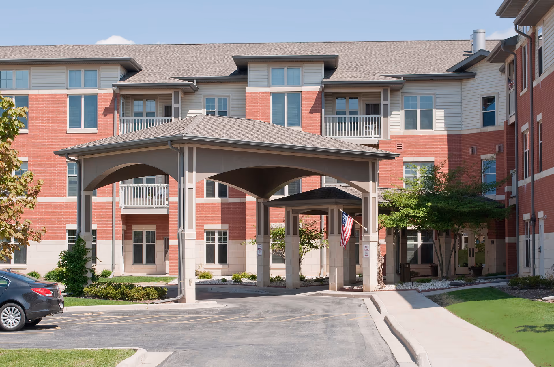 Exterior view of Dimensions Living Green Bay facility showing a covered entrance with an American flag, surrounded by a red brick and beige building with multiple windows and balconies, a parked car, and landscaped greenery.