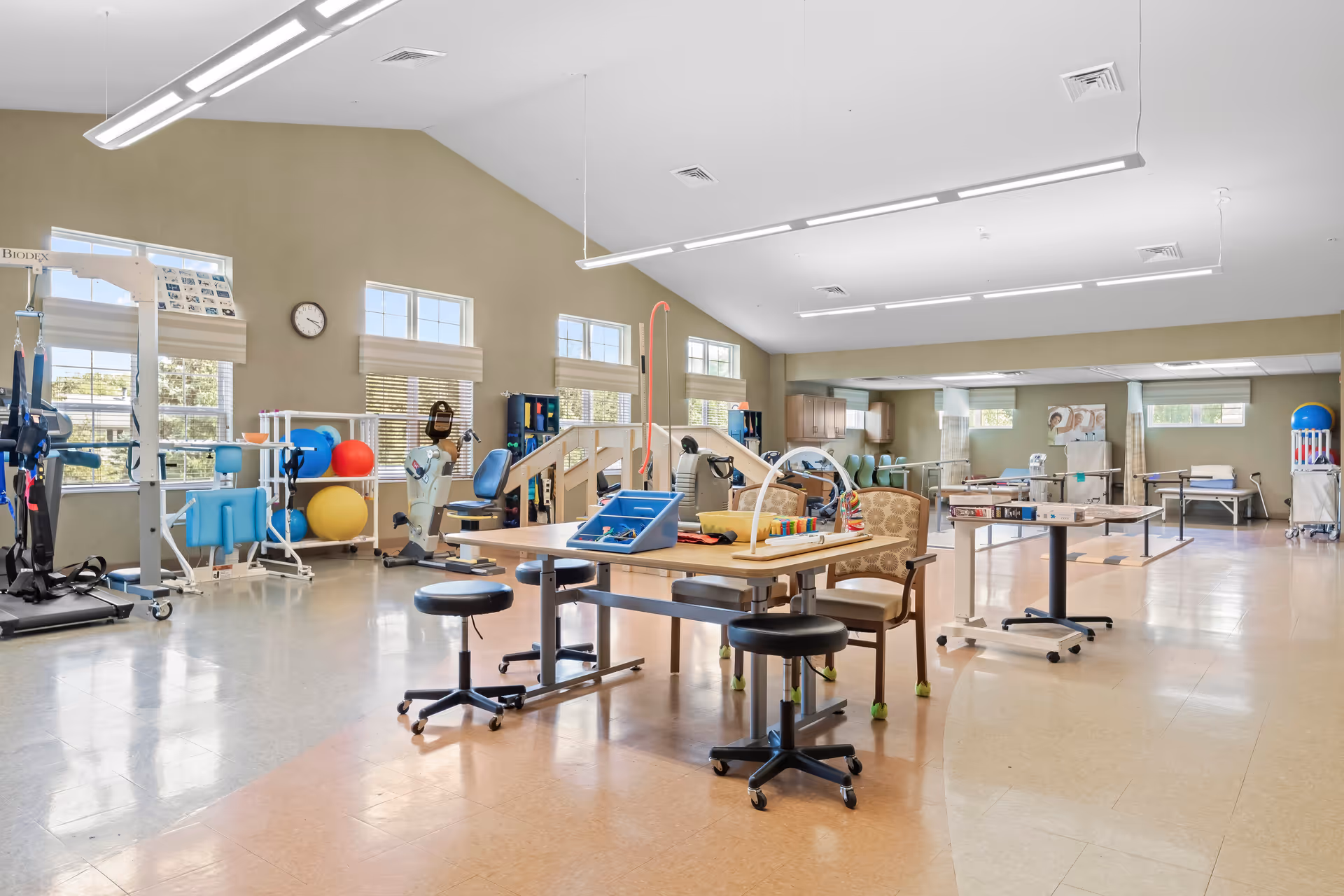 A spacious physical therapy room with exercise equipment, therapy tables, chairs, and various rehabilitation tools. The room has large windows letting in natural light and a high ceiling with hanging lights.