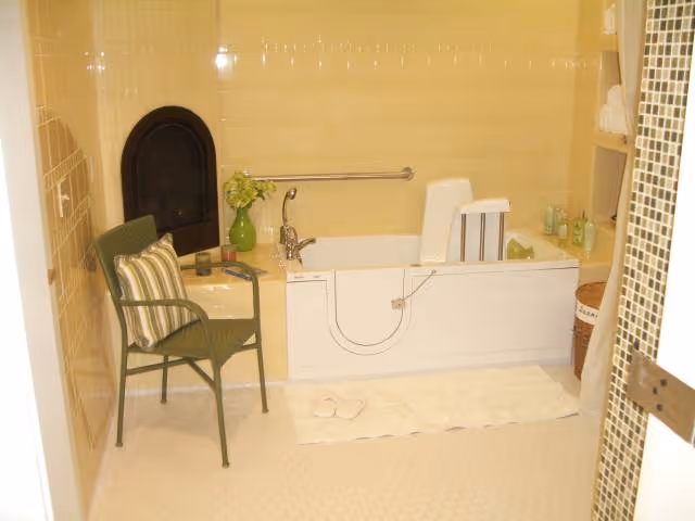 A bathroom with a walk-in bathtub equipped with a safety handle and a built-in seat. There is a green chair with a striped cushion next to the tub, a small green vase with flowers on the tub ledge, and a white bath mat with slippers on the floor. Shelves with towels and toiletries are visible on the right side.