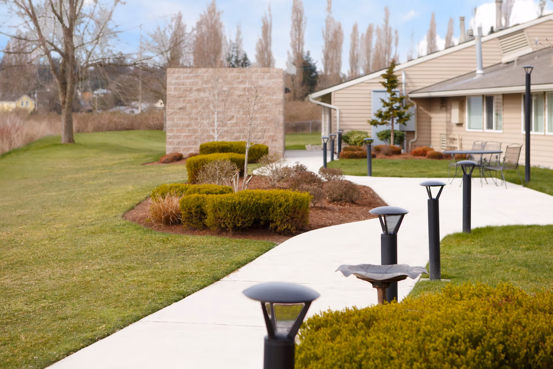 A paved walkway lined with modern black lamp posts curves through a well-maintained garden area with trimmed bushes and small trees. To the right, there is a beige building with windows and outdoor seating including tables and chairs. The background shows leafless trees and a partly cloudy sky.