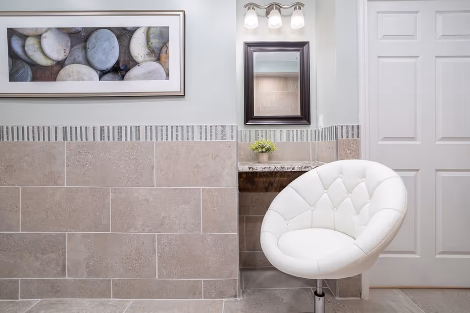 A small vanity area with a white cushioned chair in front of a granite countertop with a small potted plant. Above the countertop is a mirror with three light fixtures. The walls are tiled halfway up with beige tiles and a decorative vertical stripe border, and a framed picture of smooth stones hangs on the wall.