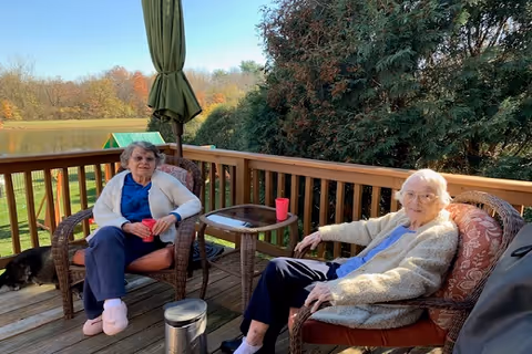 Two elderly women sitting on cushioned wicker chairs on a wooden deck with a small table between them. One woman is wearing a white cardigan and blue shirt, holding a red cup, while the other woman is wearing a beige cardigan and light blue shirt. There is a green umbrella behind them, trees in the background, and a dog lying on the deck to the left.