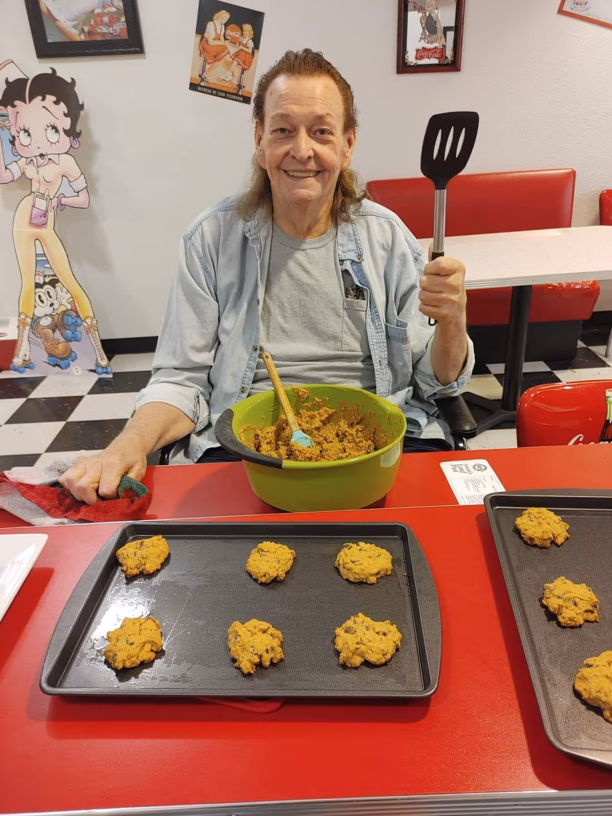 An elderly man sitting at a red table in a retro-style dining area, smiling and holding a spatula in one hand. In front of him are two baking trays with cookie dough portions and a green mixing bowl with cookie dough and a wooden spoon. The background features red booth seating and vintage-style wall art, including a large Betty Boop cutout.