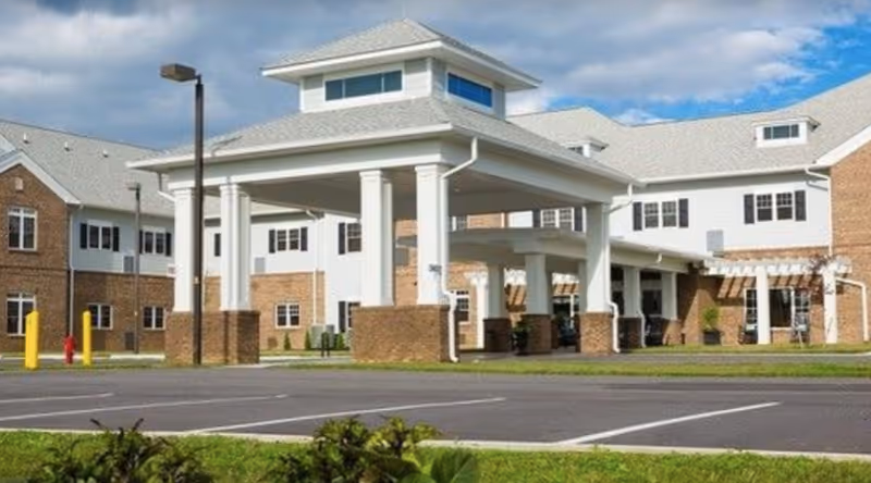 Front entrance of a senior living building with a covered porte-cochere, brick facade, and adjacent parking area.